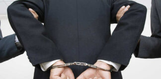 Person handcuffed in suit, escorted by two others.