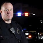 Harlem River Plunge—Cops Dive, Chaos Unfolds Police officer smiling in front of a vehicle with emergency lights