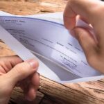 A hand holding an envelope containing a check on a wooden table