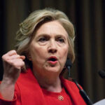 Person speaking passionately at a podium, wearing red jacket.