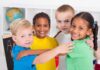 Four children of diverse backgrounds smiling and hugging each other in a classroom