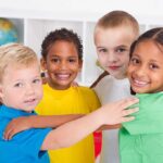 Four children of diverse backgrounds smiling and hugging each other in a classroom