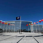 Free Ride Ends: NATO Scrambles Flags outside NATO headquarters building under clear blue sky.