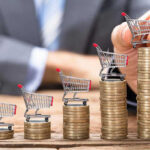 Inflation Dips, Panic Rises Mini shopping carts on stacks of coins, business background.