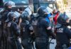 Riot police in helmets and visors stand in a line