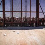 Crowd stands behind a tall metal fence at a border gate