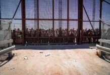 Crowd stands behind a tall metal fence at a border gate