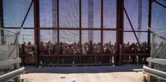 Crowd stands behind a tall metal fence at a border gate