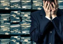 Man in suit covering face beside stacks of paperwork